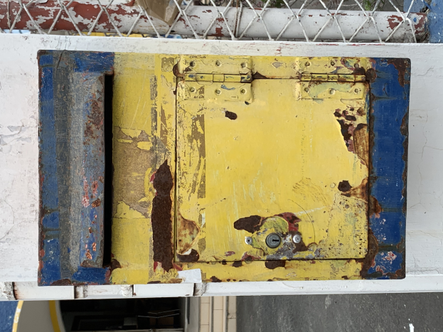 Ein gelb-blauer Metalltresor steht an einer weißen Wand, mit einem Metallzaun im Hintergrund.