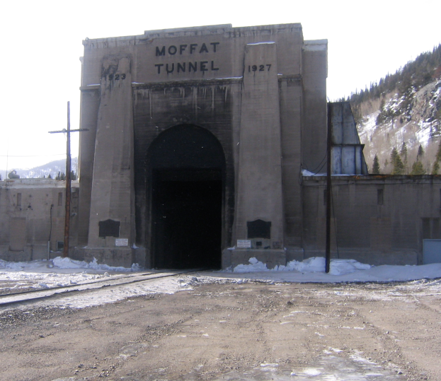 Ein Tunnel mit einem Schild, auf dem "Moffat Tunnel" steht, in der Mitte, umgeben von einer Straße, Schnee, Pfählen, Bäumen und Bergen unter einem klaren Himmel.