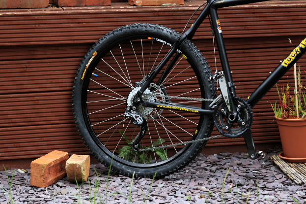 Ein Fahrrad lehnt an einer Wand mit Ziegelsteinen, Felsen und Pflanzen in der Nähe.