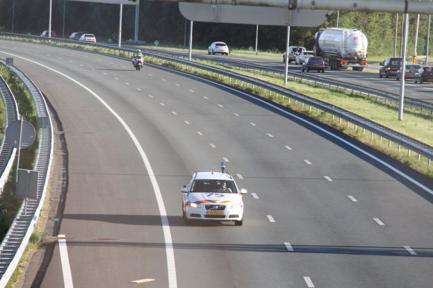 Ein Auto auf einer Autobahn mit einem stationären Polizeiwagen am Rande, andere Fahrzeuge und Bäume, Geländer, Masten und Planken im Hintergrund.