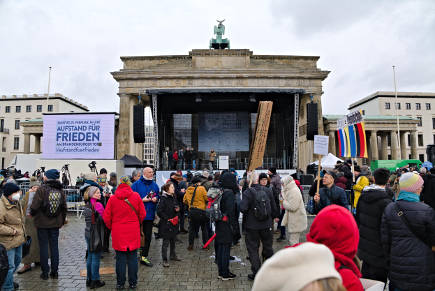 Eine Menschenmenge steht vor einem Gebäude mit einer Bühne, Lautsprechern und einem Bildschirm, mit Fahnen und Transparenten auf der rechten Seite, was auf eine Demonstration in Berlin hindeutet.
