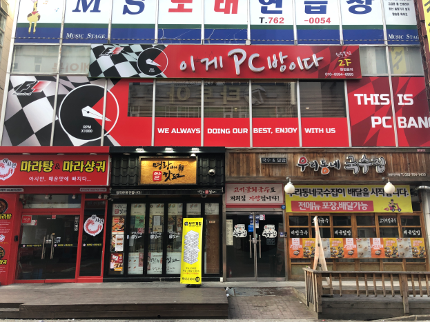 Ein modernes Gebäude mit Glaswänden und -türen, das ein Schild trägt, auf dem "Das ist die PC Bank" steht, in Seoul, Südkorea, mit einem Holzzaun davor und einer Straße unten.