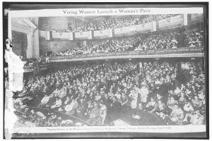 Ein Schwarz-Weiß-Foto einer großen Menschenmenge in einem Gebäude, einige sitzen, einige stehen, Banner mit Text und eine Wand im Hintergrund. Unten steht der Text "Frauen wählen eine Frauenpartei".