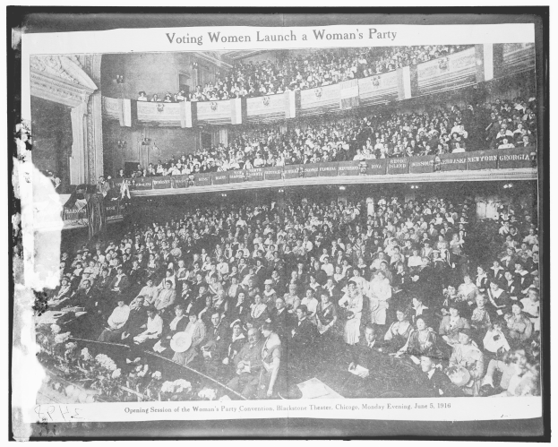 Ein Schwarz-Weiß-Foto einer großen Menschenmenge in einem Gebäude, einige sitzen, einige stehen, Banner mit Text und eine Wand im Hintergrund. Unten steht der Text "Frauen wählen eine Frauenpartei".