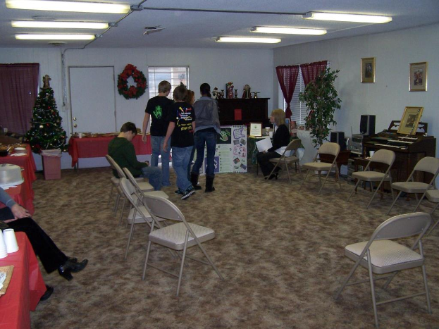 Ein Raum mit mehreren Stühlen, Tischen und Personen, einige stehen, einige sitzen, mit Tassen und Gefäßen auf den Tischen, einer Wand im Hintergrund mit einem Strauß, Vorhang, Tür, Rahmen und Fenstern, einem Weihnachtsbaum, einem Piano auf der rechten Seite und Röhrenleuchten an der Decke.