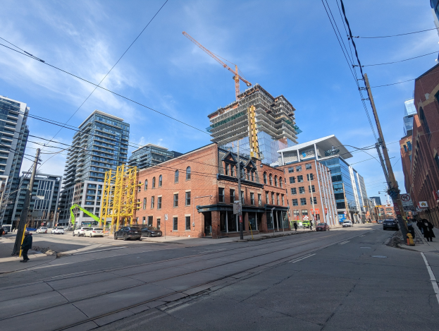 Eine belebte Stadtstraße in Toronto mit Fahrzeugen, Fußgängern, Oberleitungen, Schildern und Gebäuden, darunter eine Baustelle mit einem Kran im Hintergrund vor einem bewölkten Himmel.