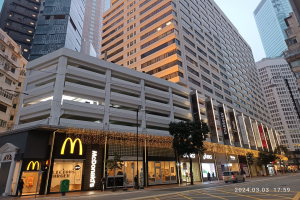 Ein McDonald's-Restaurant auf einer belebten Stadtstraße, flankiert von hohen Gebäuden, Straßenlaternen und Verkehr, mit Fußgängern auf dem Gehweg unter einem klaren blauen Himmel.