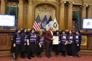 Eine Gruppe von Menschen steht vor Flaggen, ein Mann in der Mitte hält ein Zertifikat, flankiert von zwei Bildschirmen und einer hölzernen Wand mit kunstvollen Schnitzereien und Vorhängen im Hintergrund, wahrscheinlich eine Stadtratssitzung.