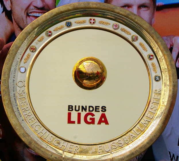Goldener Schild mit "Bundesliga"-Text, gehalten von einem Mann, mit einem Banner im Hintergrund, das drei Personen zeigt.
