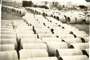 Ein Schwarz-Weiß-Foto eines großen Haufens von Betonrohren auf dem Boden, mit ein paar Gebäuden im Hintergrund.