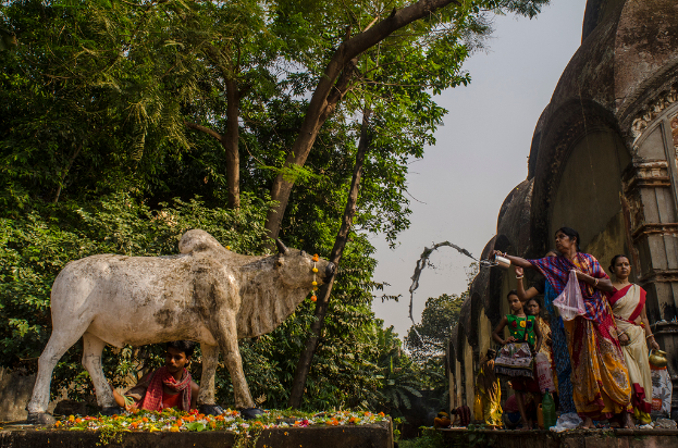 Eine Kuhskulptur mit einem Mann dahinter, umgeben von Blumen, mit ein paar Menschen neben einem Tempel und Bäumen im Hintergrund.