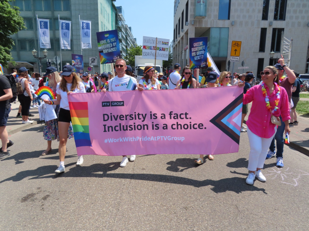 Eine Gruppe von Menschen marschiert auf einer Stra├če, h├Ąlt ein Banner mit der Aufschrift "Diversit├Ąt ist eine Tatsache, Inklusion ist eine Wahl", einige tragen Mützen und Schutzbrillen, andere halten weitere Banner, vor einem Hintergrund aus Geb├Ąuden, B├Ąumen, Laternenmasten und einem klaren blauen Himmel.