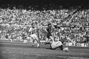 Schwarzes Foto von Männern, die auf einem Feld Füßball spielen, mit Zuschauern im Hintergrund und einer Fahne mit der Aufschrift "Bundesliga 1978-79 Ajax 1-1 Hannover 96."