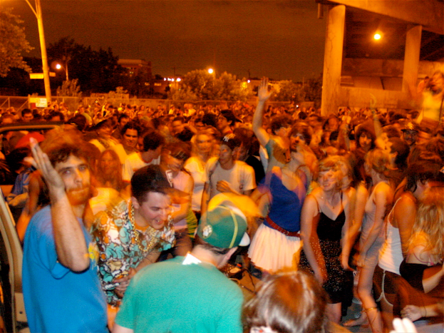 Menschen tanzen vorne, während im Hintergrund Lichter, Stangen, Bäume und andere Gegenstände zu sehen sind.