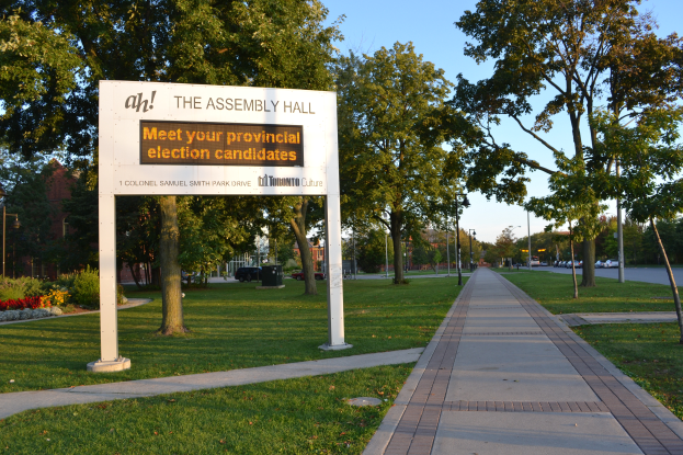 Ein Schild auf dem Boden mit der Aufschrift "Die Versammlungshalle - Treffen Sie Ihre Landtagswahlkandidaten" umgeben von Gras, einem Weg, Bäumen, blühenden Pflanzen, Straßenlaternen, Fahrzeugen, einem Gebäude mit Fenstern und einem bewölkten Himmel.