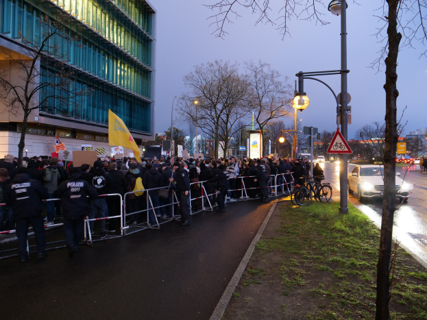 Große Gruppe von Menschen vor einem Gebäude mit Barrikaden, Schildern und Bäumen, was auf eine Demonstration in Berlin hinweist.