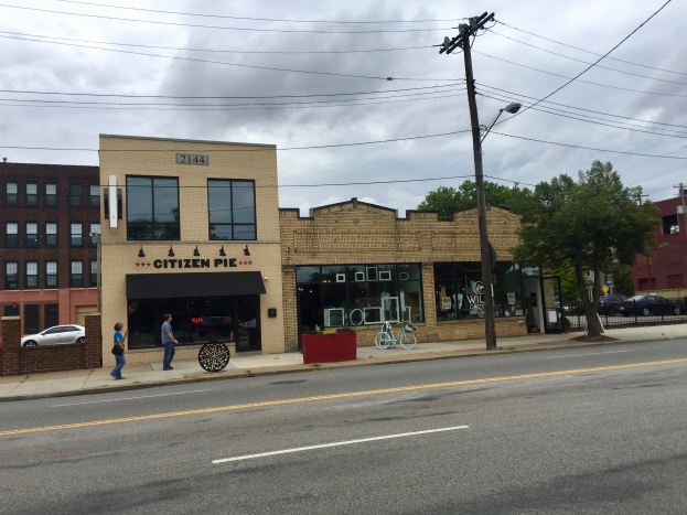 Eine belebte Stadtstraße mit Fußgängern, parkenden Fahrrädern, Strommasten, Bäumen und Gebäuden, mit dem Citizen Pie Restaurant an der Ecke.