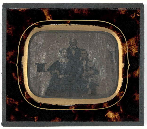 Ein altes Foto eines Mannes im Anzug und einer Frau in unbekannter Kleidung vor einem Fernseher stehend, beide mit ernsten Gesichtern.