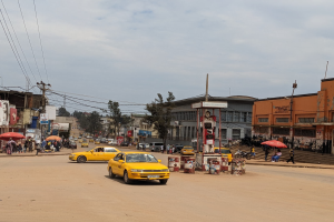 Eine belebte Stadtstra├če mit einem gelben Taxi, das f├Ąhrt, Passanten mit Schirmen auf den Gehwegen, Geb├Ąude mit Fenstern, Strommasten mit Kabeln und ein bew├Âlktes Himmel.