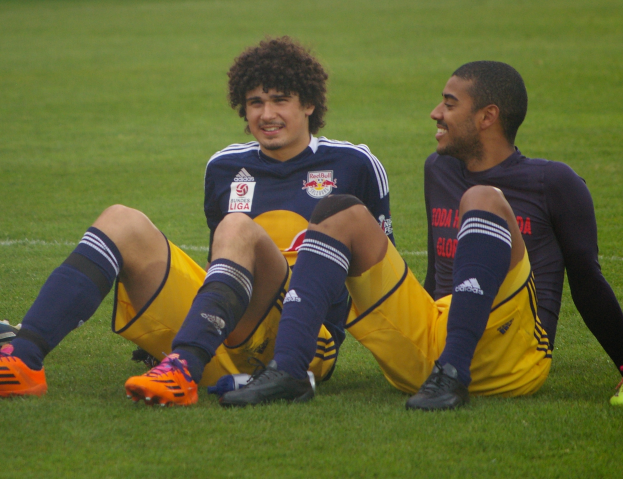 Zwei Fußballspieler sitzen auf dem Boden mit gekreuzten Beinen, tragen Sportbekleidung und lächeln sich an.