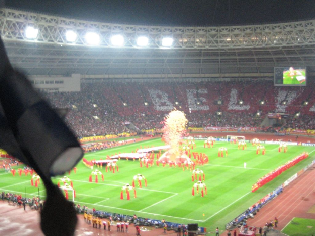 Ein Stadion mit Flutlicht, Eisengittern, Zuschauern, stehenden Individuen, Werbetafeln, Anzeigetafeln, Sicherheitskameras, Flaggen und Deko.