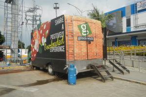 Lastwagen vor einem Gebäude mit einer Tafel, auf der 'Teilen Sie eine positiv überladene Erfahrung' steht, mit Treppen, einem Zylinder auf dem Boden, Geländern, Strom- und Lichtmasten, Bäumen und einem bewölkten Himmel.