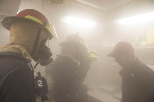 Feuerwehrleute mit Helmen und Rauch aus dem Mund, einer hält eine Kamera, während einer Trainingsrunde mit Hintergrundbeleuchtung und -gegenständen.