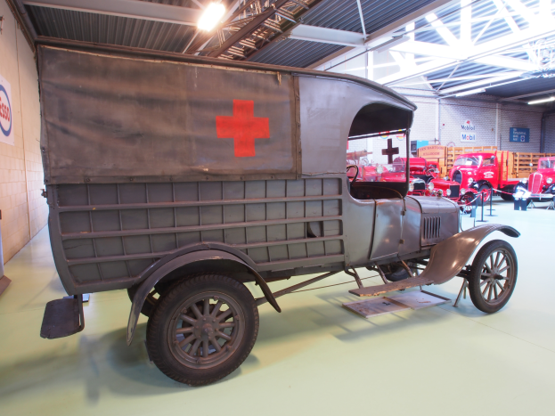 Ein alter Krankenwagen mit einem roten Kreuz auf der Seite steht in einem Gebäude geparkt, umgeben von anderen Fahrzeugen, mit einer Texttafel an der linken Wand und Lampen auf dem Dach.