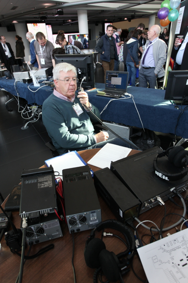 Ein älterer Mann sitzt auf einem Stuhl und hält ein Mikrofon in der Hand, während verschiedene elektronische Geräte auf dem Tisch vor ihm liegen und eine Gruppe von Menschen hinter ihm steht und die Geräte ansieht, unter einer Decke mit Spotlights.