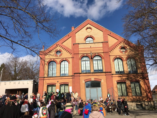 Eine große Gruppe von Menschen, die vor einem mehrstöckigen Gebäude mit Fenstern und Türen stehen, umgeben von Bäumen unter einem bewölkten Himmel, möglicherweise an einem Festival teilnehmen.