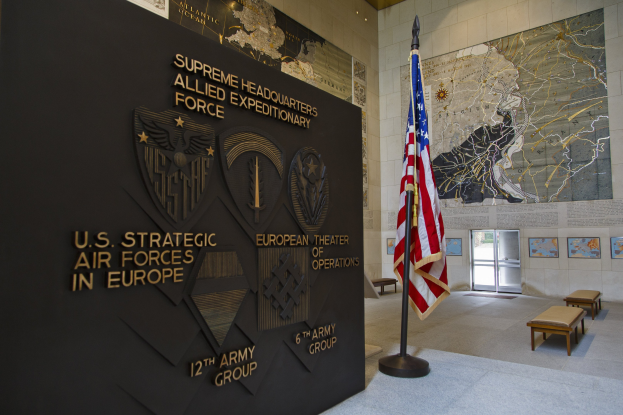 Eingang zum US-Strategischen Luftstreitkräfte-Hauptquartier in Europa mit einer amerikanischen Flagge, Text und Logos an der Wand, Bänken, einer Tafel, Fotorahmen, beleuchteter Decke und einer Tür im Hintergrund.