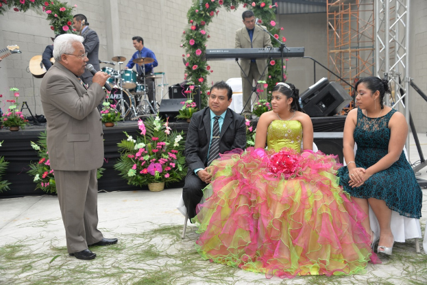 Ein Mann steht an einem Mikrofon und spricht, während im Hintergrund ein Orchester spielt und die Bühne mit Blumen und Pflanzen geschmückt ist, mit einigen Menschen, die sitzen und anderen, die stehen.