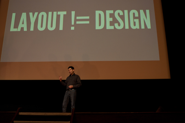 Eine Person steht auf einer Bühne und spricht, im Hintergrund ist ein Bildschirm mit Text zu sehen.