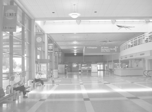 Ein Schwarz-Weiß-Bild eines Innenraums mit zwei Personen auf einer Bank links, einem Schreibtisch rechts, einem Eingang in der Mitte, Richtungsschildern oben und einer Rolltreppe rechts mit einigen Personen vor einem Zaun sitzend.