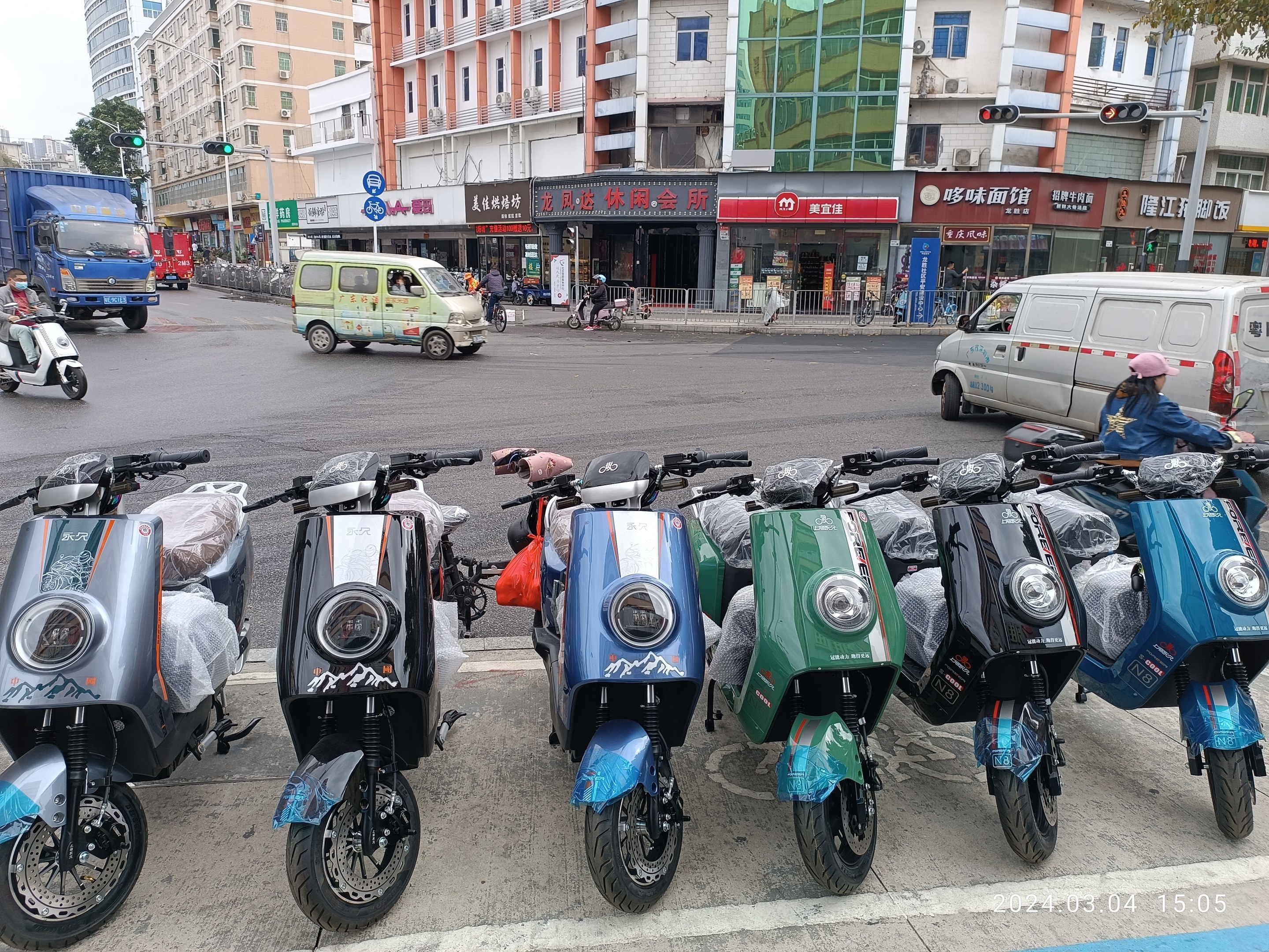 Eine Reihe von Elektro-Scootern, die auf einer Straße geparkt sind, mit einer Person in einer Mötte davor, Gebäude, Schilder, Verkehrszeichen, Bäume und einen klaren blauen Himmel im Hintergrund.