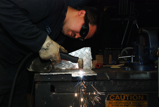 Ein Schweißer in Schutzbrille und Handschuhen bedient eine Schweißmaschine an einem Metallteil, mit einem Warnschild und verstreuten Werkzeugen im Hintergrund.