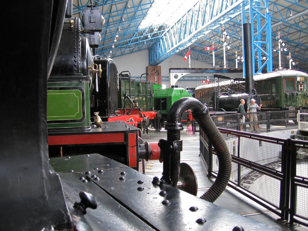 Mehrere Lokomotiven stehen in einem Abstellgleis mit zahlreichen Eisenstangen darüber. Rechts verläuft ein Zaun entlang einer Eisenbahnschiene mit einem Zug. Zwei Personen stehen in der Mitte der Szene nahe dem Zaun.