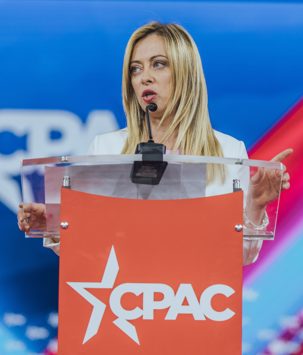 Eine Frau in einem weißen Kleid steht an einem Rednerpult mit einem Mikrofon, sieht aus wie eine Politikwissenschaftlerin.