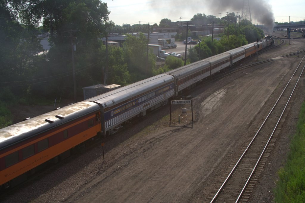 Ein Zug auf Schienen mit Bäumen, Strommasten mit Kabeln, Autos und Gebäuden links, eine Brücke vorne und Rauch aus dem Zug.