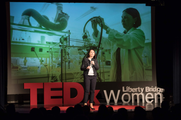 Frau auf einer Bühne mit Mikrofon stehend, mit einem Bildschirm dahinter, der ein Bild einer Frau und Text zeigt, der sich auf TEDxWomen bezieht.