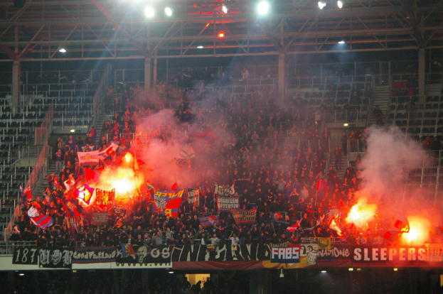 Eine große Menge Menschen in einem Stadion hält Fahnen und Banner, mit Rauchwerfern, unter einem Dach mit Deckenlampen und Metallrahmen.