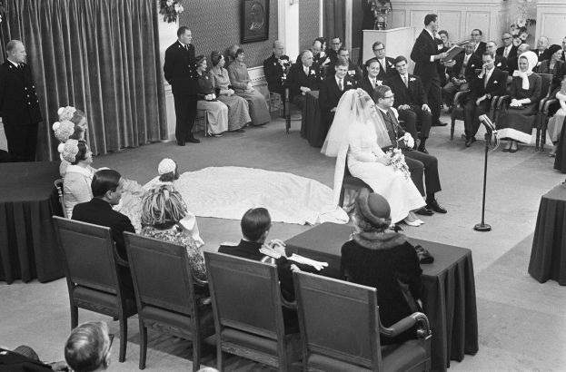 Schwarzes und weißes Foto einer Hochzeitszeremonie mit Menschen, die sitzen und stehen, einem Mann mit Mikrofon, einer Frau mit einem Blumenstrauß, einem Podium mit Mikrofon, Blumenschmuck, einem Vorhang, einem Rahmen an der Wand und einer Tür im Hintergrund.