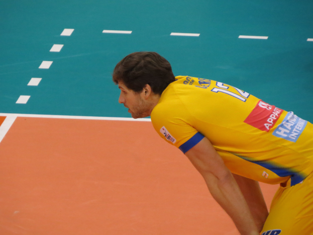 Eine professionelle Volleyballspielerin in einem gelben 'Volleyball'-T-Shirt steht bereit, um auf einem Volleyballfeld zu dienen.