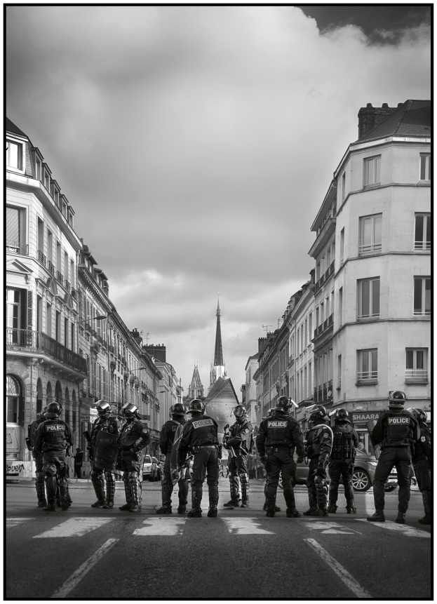 Eine Gruppe von Polizisten in Helmen steht in der Mitte einer Straße umgeben von Fahrzeugen, mit Gebäuden auf beiden Seiten und einem bewölkten Himmel im Hintergrund; Text ist unten im Bild sichtbar.