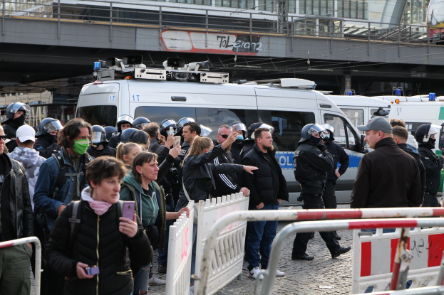 Eine Gruppe von Menschen steht vor einer Reihe von Polizeifahrzeugen, einige tragen Helme und halten Mobiltelefone, mit Barrikaden im Vordergrund und einer Brücke mit Geländern und Gebäuden im Hintergrund in Berlin, Deutschland.