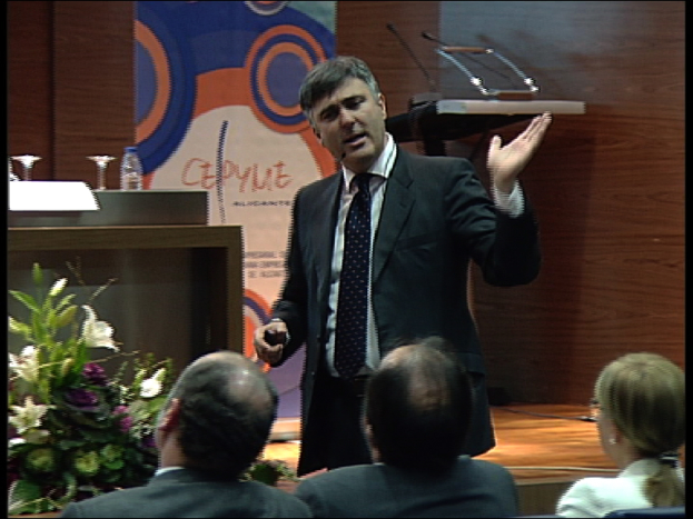 Ein Mann in einem schwarzen Blazer steht an einem Podium mit Mikrofonen, spricht in ein Mikrofon, mit einem Blumentopf oder einem Blumenstrauß daneben und einem Tisch mit einem Buch, einer Wasserflasche und anderen Gegenständen daneben, während drei Personen auf Stühlen vor ihm sitzen, mit einer braunen Wand und einer bunten Tafel mit Text im Hintergrund, wahrscheinlich in einem Konferenzsaal.