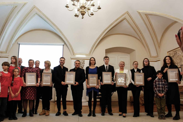 Gruppe von Menschen mit Urkunden bei einer Preisverleihung mit einer Wand, einer Tür, einem Bildschirm und einem Kronleuchter im Hintergrund.