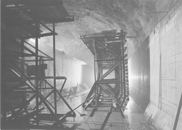 Schwarz-weiß-Foto eines Tunnels mit einer zentralen Metallstruktur, Metallstangen auf der linken Seite, einer Wand auf der rechten Seite und Text unten.