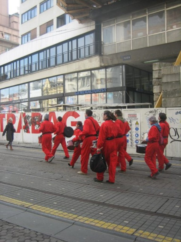 Menschen, die auf einem Boden gehen und Musikinstrumente spielen, mit einer Umzäunung, die Plakate und Text zeigt, im Hintergrund und Gebäude mit Glaswänden und Fenstern weiter hinten, einige mit Namen darauf.