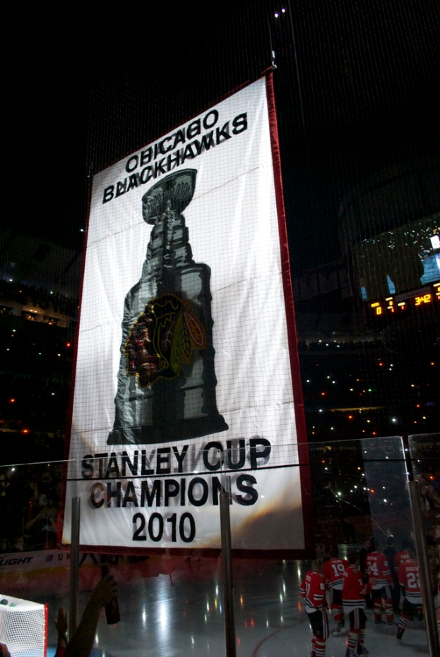 Eine nächtliche Stadionszene mit einem großen Banner in der Mitte, Zuschauern im Hintergrund, einem Glas auf dem Boden, ein paar Spielern auf dem Feld und beleuchteten Rängen.
